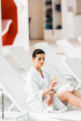 attractive woman holding glass of champagne and sitting in bathrobe on deck chair