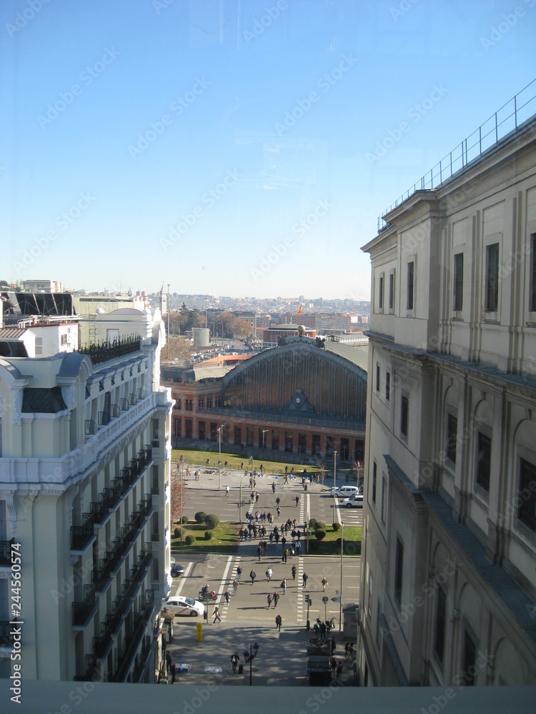 Fototapeta premium 2ª vista urbana desde altura con primer plano de edificios históricos y detrás con calzada, coches, antigua estación de tren y horizonte de fondo ciudad con nuves y cielo azul