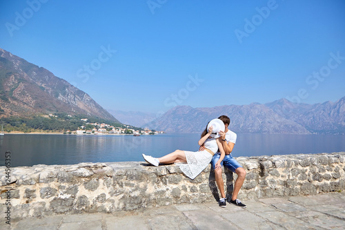 Cute couple in love kissing behind white female hat , sitting on the stones on the background of the sea coast, mountains and blue sky © mikeosphoto