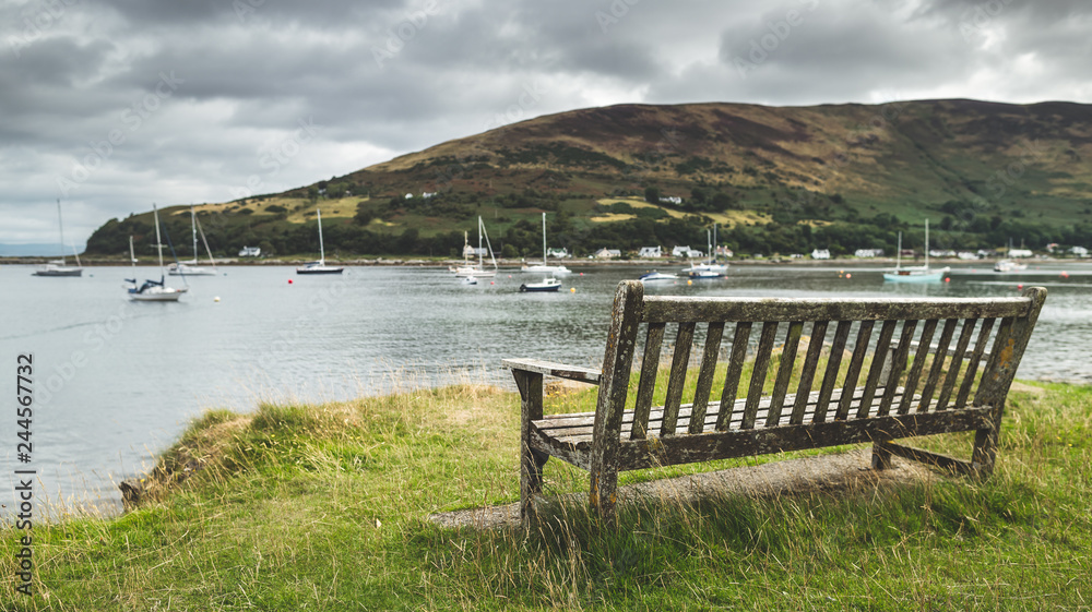 Close-up bench on the Arran island shore. Amazing bayview. Breathtaking Scottish landscape. Cozy bay with the yachts under the rainy cloudy sky. Ideal place for the rest and relax.