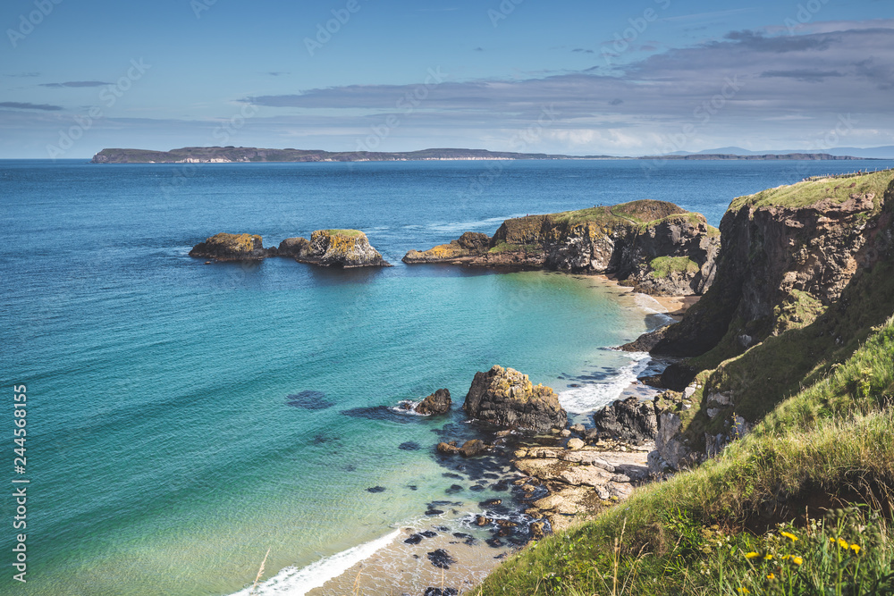 Sunlit turquoise ocean water washing Northern Ireland shore. Stunning marine scene. The grass covered cliffs next to the cozy white sand beach. The Irish landscape with the cloudy sky background.