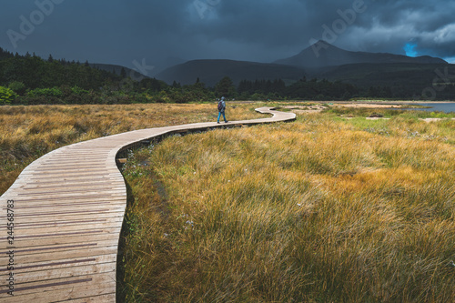 Tourist walking on the wooden path. Northern Ireland. Mighty mountains background. Rainy cloudy sky in grey tints. The backpacker on the trail. Hiking among the wild environment. Outdoor activity.
