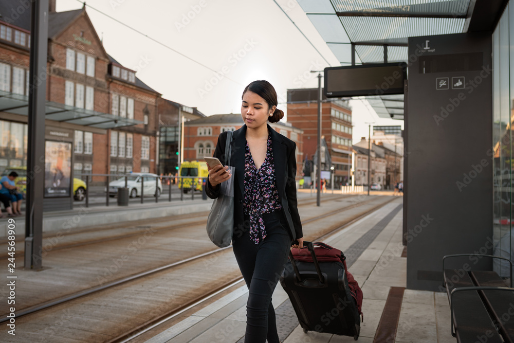 Asian businesswoman waiting with her luggage using a cellphone - obrazy, fototapety, plakaty 