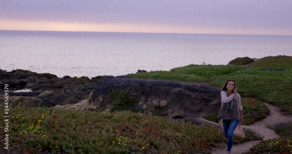 Young happy woman brushes hair from face as she walks along path with ocean in the background Young happy woman brushes hair from face as she walks along path with ocean in the background
