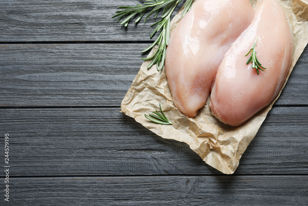 Parchment with raw chicken breasts and rosemary on wooden background, top view. Space for text