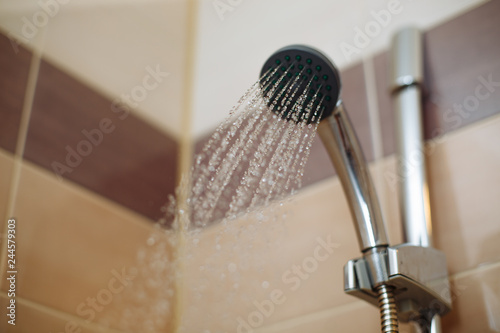  Photo of a shower head with flowing water. Water pours out of the shower, the walls are decorated with coffee-colored tiles.