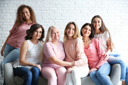 Group of women with silk ribbons sitting on sofa against brick wall. Breast cancer awareness concept