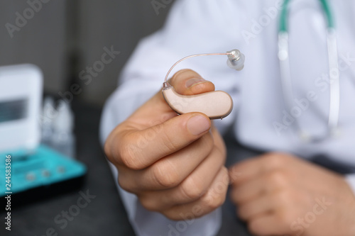 Doctor holding hearing aid against blurred background, closeup. Medical objects