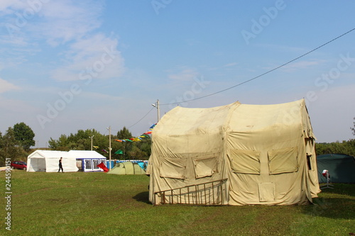Large sandy canvas tent on the green grass near the trees on a blue sky background on summer day - sports tourism, camping, scouting © Ilya