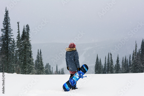 snowboarder girl stand on top of a mountain turning back to camera holding her snowboard and watching downhill of a mountain. Prepearing to ride and searching for her line. Freeride in blizzard.