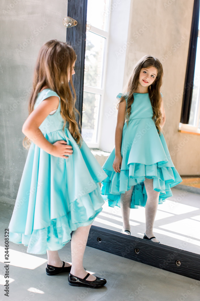 The lovely little girl posing in a beautiful blue dress is posing looking at the mirror
