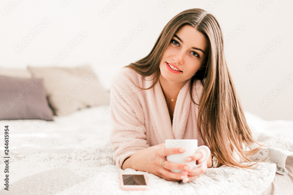 Young woman lying in white bed and using a phone in her bedroom. Happy morning. Young woman lying in white bed and using a phone in her bedroom. Happy morning.