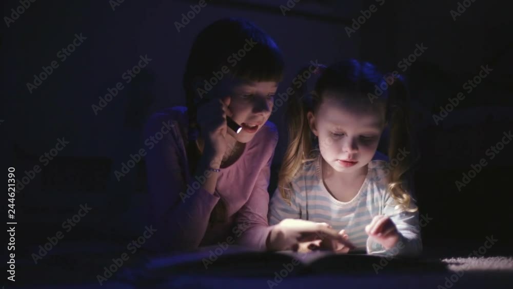 two sisters reading a book with a flashlight in a dark room at night two sisters reading a book with a flashlight in a dark room at night