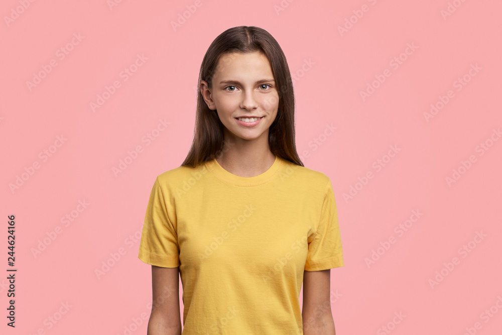 Photo of pretty teen girl has serious look at camera, long hair, has gentle smile, shows her natural beauty. Teenager poses against pink background with copy space for advertisement Photo of pretty teen girl has serious look at camera, long hair, has gentle smile, shows her natural beauty. Teenager poses against pink background with copy space for advertisement