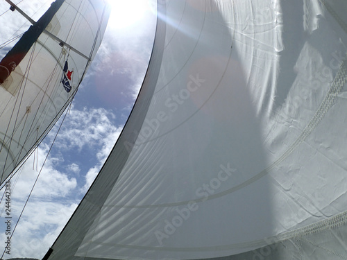 Looking up through full white sails with a French and a Martinique courtesy flag flying in the rigging, and a burst of sunlight at the top in a blue sky full of puffy clouds. photo