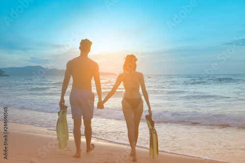 Happy Romantic Young Asian Couple Enjoying Beautiful Sunset Walk on the Beach. Cheerful couple Holding With Mask And Flippers For Snorkel On Sunset Time. Travel Vacation Lifestyle Concept. 