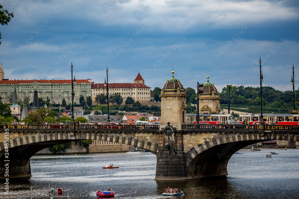 Fototapeta premium PRAGUE, CZECH REPUBLIC - AUGUST 28, 2015: Pedal boats sail and pass under the medieval stone Chec Bridge on Vltava River, Prague, Czech Republic in calm summer evening with people walking on top