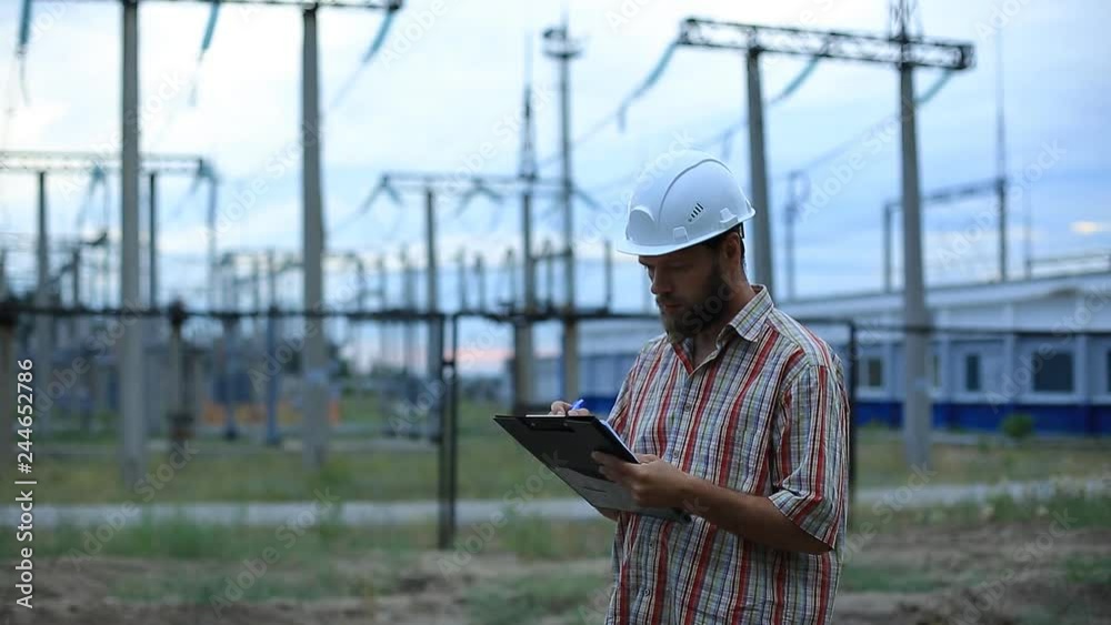 Station technical director with working drawings at nuclear power station. Worker in white helmet with engineering drawing near outdoor switchgear at nuclear power station Station technical director with working drawings at nuclear power station. Worker in white helmet with engineering drawing near outdoor switchgear at nuclear power station