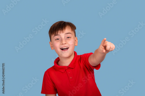 Waist up portrait of a smiling boy wearing a red shirt pulling a hand forward pointing to something on blue background in studio