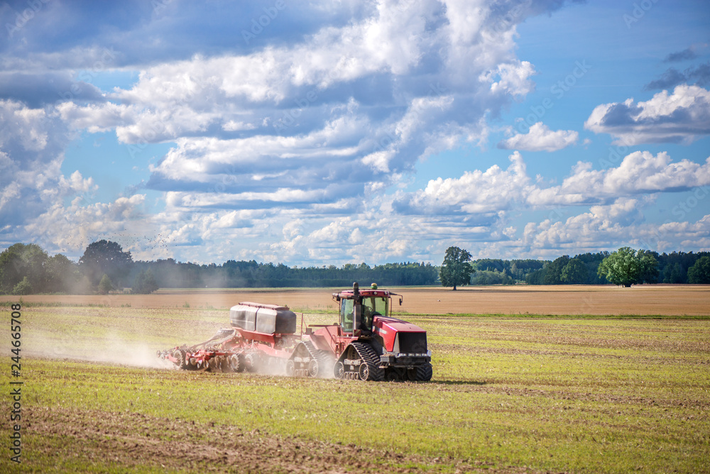 Fototapeta premium Agricultural background with red tractor pulling plow, throwing dust in air. Combine harvester at wheat field. Heavy machinery during cultivation, working on fields. Dramatic sky, rain, storm clouds