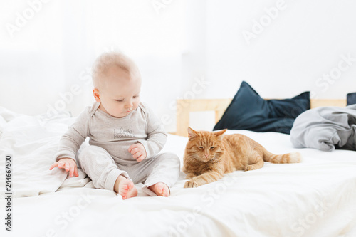 A small cute baby sits next to a ginger cat on the bed in a cozy home setting