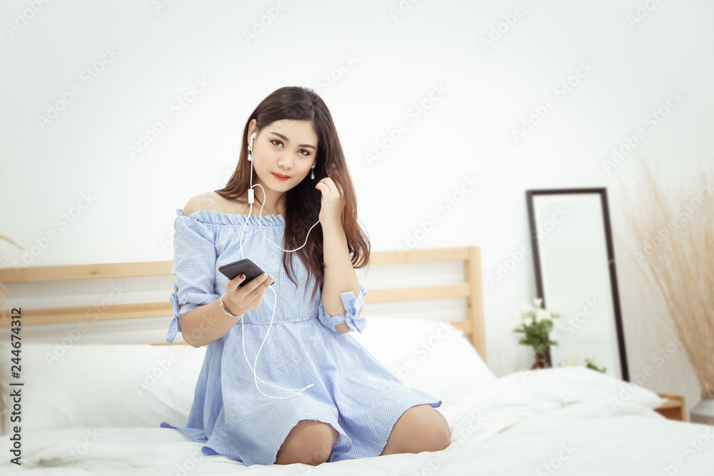 A beautiful Asian woman with headphone relaxing at home , sitting on the bed listening to music from application on mobile phone A beautiful Asian woman with headphone relaxing at home , sitting on the bed listening to music from application on mobile phone