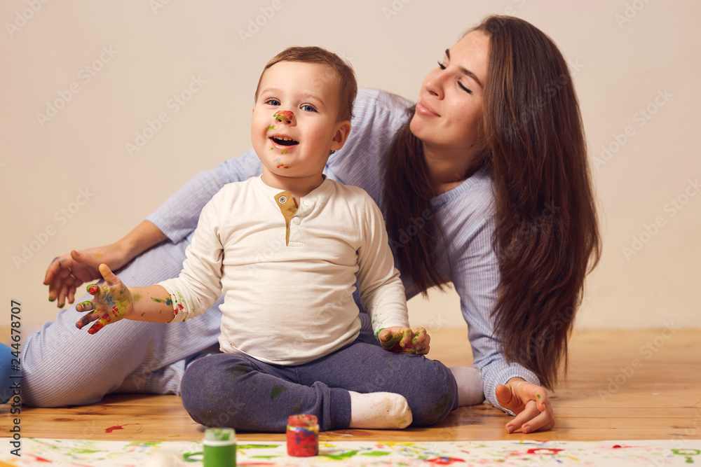 Naklejka premium Happy mother and her little son with paints on his face dressed in home clothes are sitting on the wooden floor in the room and painting with fingers on the white paper