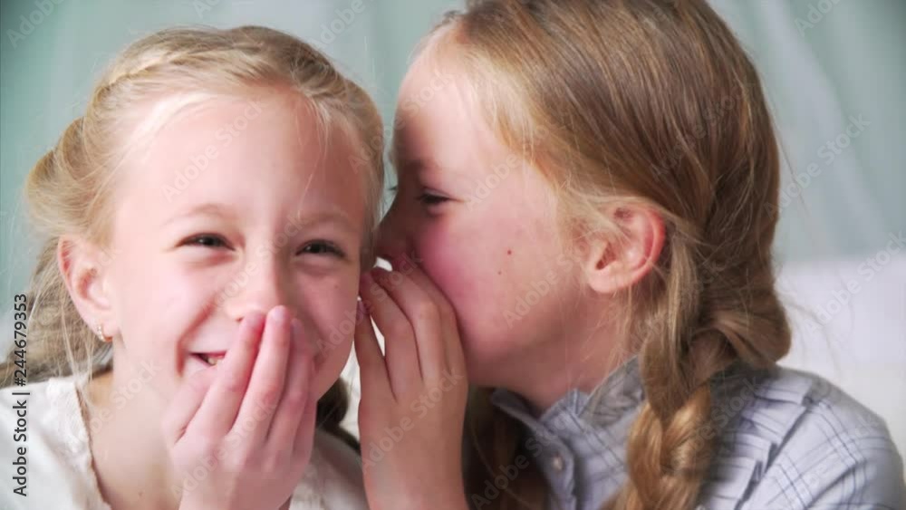 Portrait of two adorable girls with long braids. One is whispering something in her sister's ear, they are giggling and having fun Portrait of two adorable girls with long braids. One is whispering something in her sister's ear, they are giggling and having fun