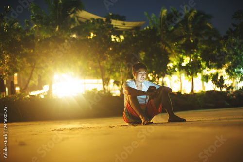 asian man using mobile and sitting on the sandy beach at night alone
