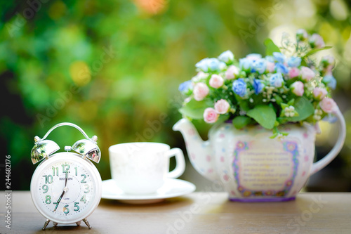 Alarm clock  with a cup of coffee and flower pots background in the morning sunlight.