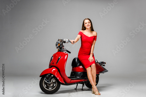 beautiful girl in red dress looking at camera and posing while sitting on motor scooter on grey background
