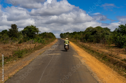 Typical road in Middle Vietnam with rider on motorbike. On the road are holes. Along the road are forest and red clay. Cloudy weather and sunshine. Near village Pleiku.