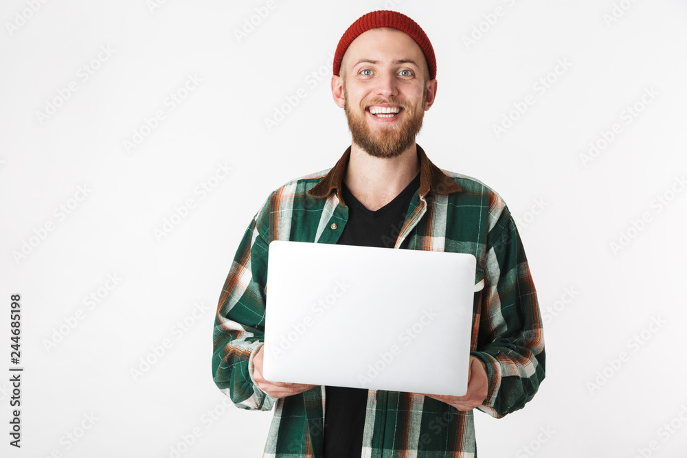 Portrait of cheerful guy wearing hat and plaid shirt holding silver laptop, while standing isolated over white background Portrait of cheerful guy wearing hat and plaid shirt holding silver laptop, while standing isolated over white background