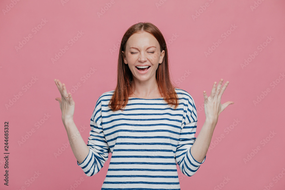Waist up shot of overjoyed pretty woman gestures with hands, laughs positively, being in high spirit, dressed in sailor striped jumper, closes eyes shut, poses against pink studio wall. Happiness Waist up shot of overjoyed pretty woman gestures with hands, laughs positively, being in high spirit, dressed in sailor striped jumper, closes eyes shut, poses against pink studio wall. Happiness