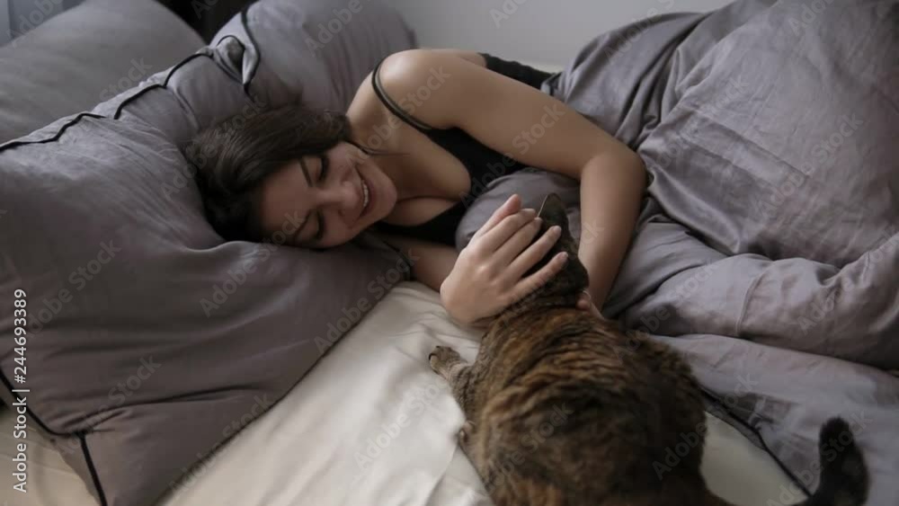 Pets, comfort, rest and people concept - happy young woman with cat lying in bed at home. Brunette girl relaxed caresses her cat in the bed Pets, comfort, rest and people concept - happy young woman with cat lying in bed at home. Brunette girl relaxed caresses her cat in the bed