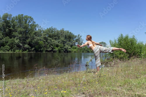 Red-haired girl doing yoga on the river bank on a background of trees on a summer sunny day.