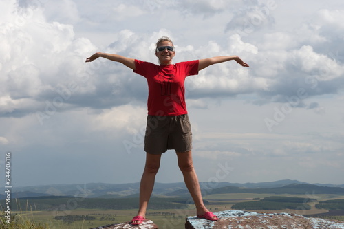 Fotografie An adult woman stands, arms outstretched, on a high mountain, against the backdrop of a plain and a cloudy sky