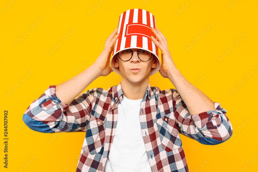 man in glasses is happy to watch a movie, with a bucket of popcorn on his head, on a yellow background man in glasses is happy to watch a movie, with a bucket of popcorn on his head, on a yellow background