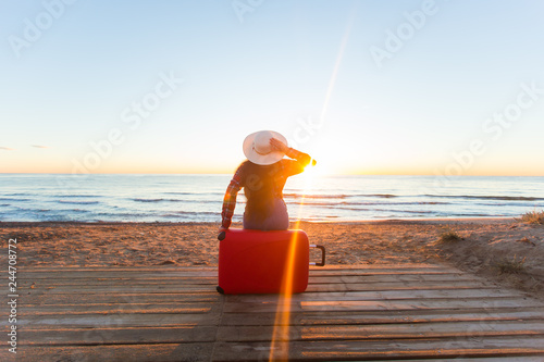 Travel, holiday and people concept - woman tourist sitting near the sea on the red suitcase and in hat and watching sunset