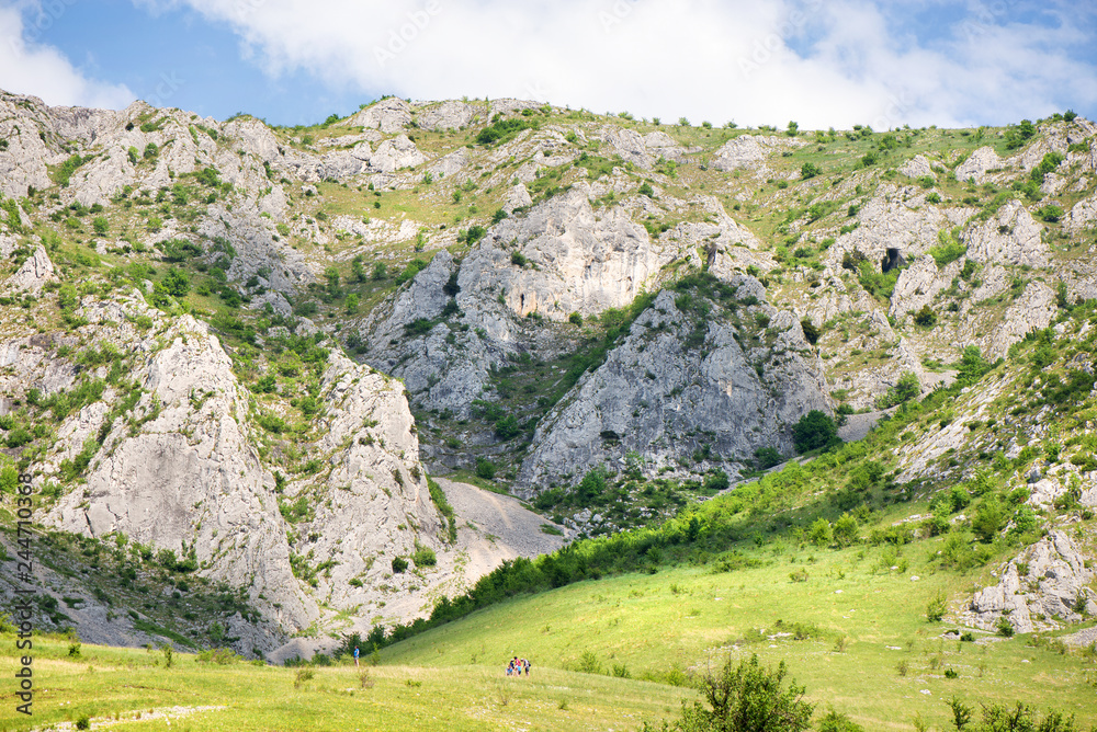 Obraz premium Sunny summer day with white clouds over a mountain peak, where tourists climb to conquer fear, find courage and develop lateral thinking skills to overcome the difficulty of climbing a mountain. 