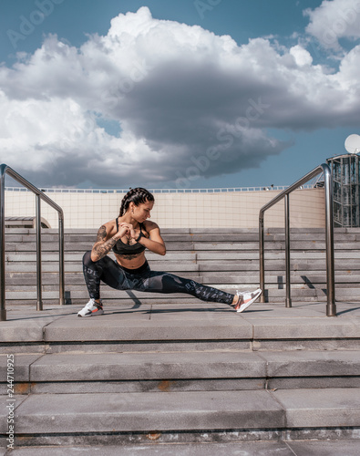Girl athlete does stretching, before jogging in morning in the city on the steps. Against the backdrop of concrete and clouds. Healthy lifestyle. In sportswear and sneakers. Tanned skin and tattoos.
