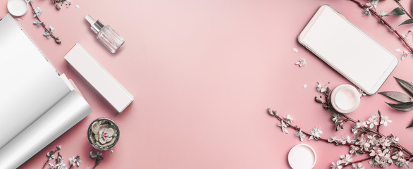 Smartphone and open magazine mock up on pastel pink desktop background with cosmetics and blossom branches, top view. Beauty blog and female business concept. Flat lay frame . Banner or template