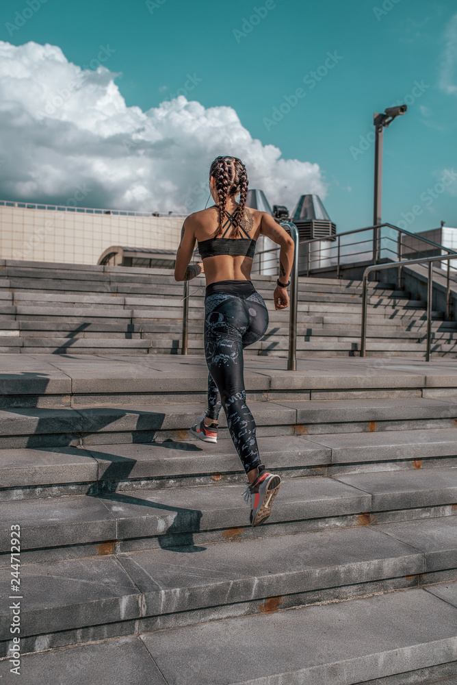 The girl athlete walks up the stairs, runs for a morning jog. In the summer in the city. The concept of a healthy lifestyle. View from the rear on a background of blue sky and concrete stairs.