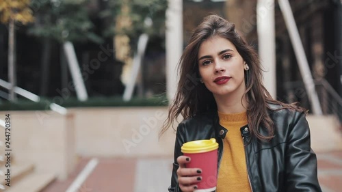 Happy young woman walking near the business center and dancing with a cup of coffee in hand. Beautiful girl looking into the camera. Good mood, funny, slow motion photo