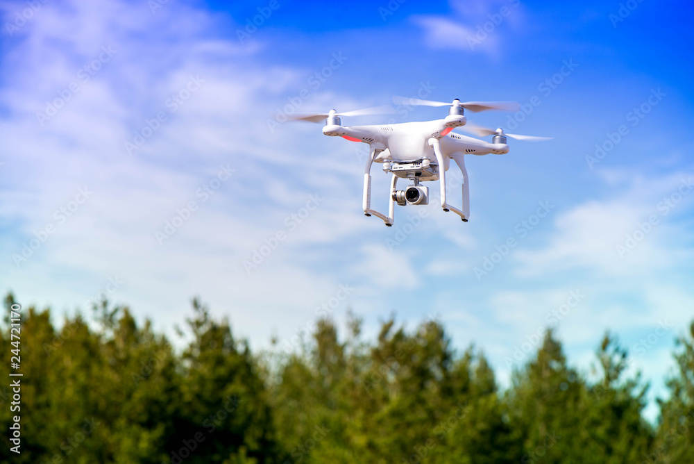 White quadrocopter is flying high in the air, taking photos and recording footage from above. Flying drone with four motors and propellers, camera and red warning lights on clear blue sky background. White quadrocopter is flying high in the air, taking photos and recording footage from above. Flying drone with four motors and propellers, camera and red warning lights on clear blue sky background.