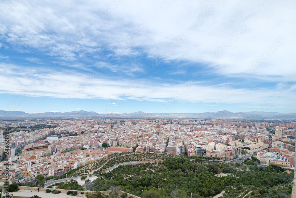 Fototapeta premium Panoramic view of city from Santa Barbara Castle in Alicante, Spain. Block apartment buildings, parks, roads, houses, palm trees. Beautiful mountain landscape in background, blue sky 