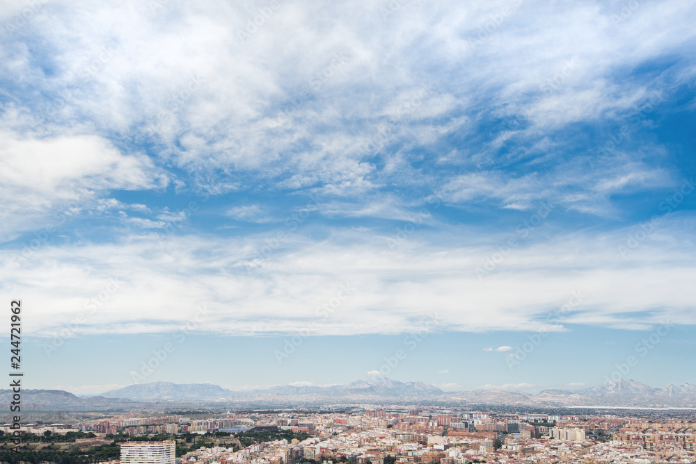 Panoramic view of city from Santa Barbara Castle in Alicante, Spain. Block apartment buildings, parks, roads, houses, palm trees. Beautiful mountain landscape in background, blue sky  