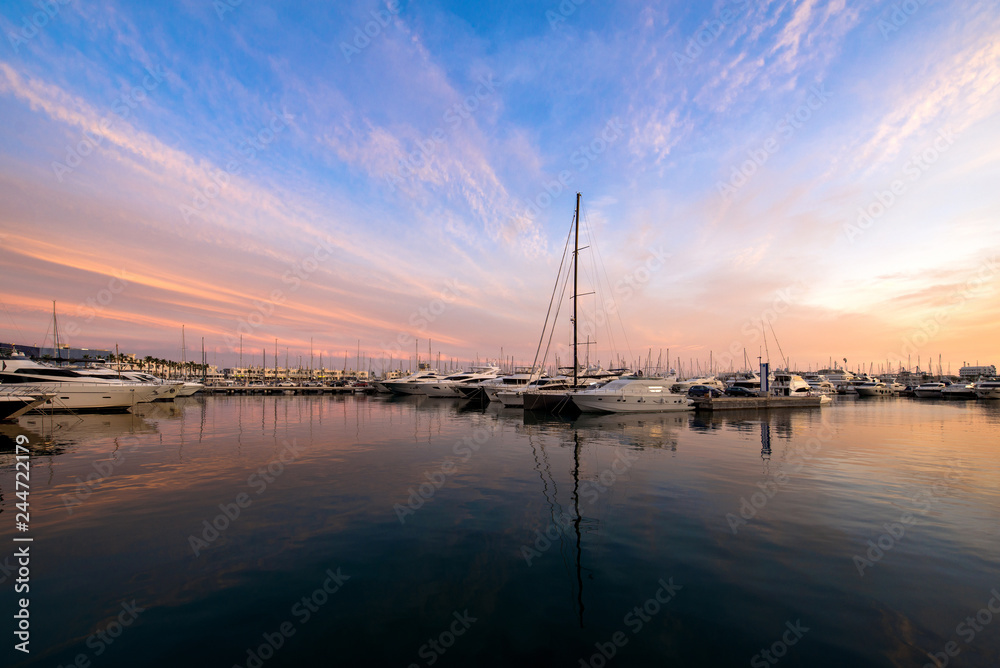 Beautiful port of Alicante, Spain at Mediterranean sea. Luxury yachts, ships, ferries and fishing boats sailing and standing in rows in harbor. Rich people traveling around the world. Sunset evening  