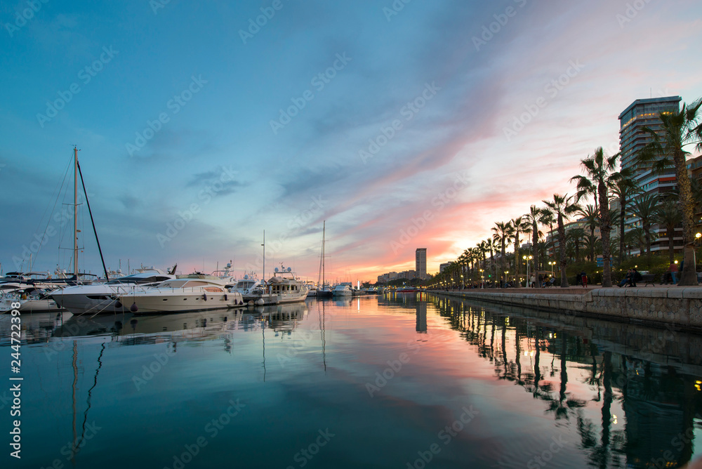 Beautiful port of Alicante, Spain at Mediterranean sea. Luxury yachts, ships, ferries and fishing boats sailing and standing in rows in harbor. Rich people traveling around the world. Sunset evening  