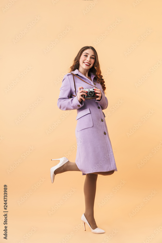Full length photo of elegant woman 20s with long hair smiling and holding retro camera, standing isolated over beige background Full length photo of elegant woman 20s with long hair smiling and holding retro camera, standing isolated over beige background